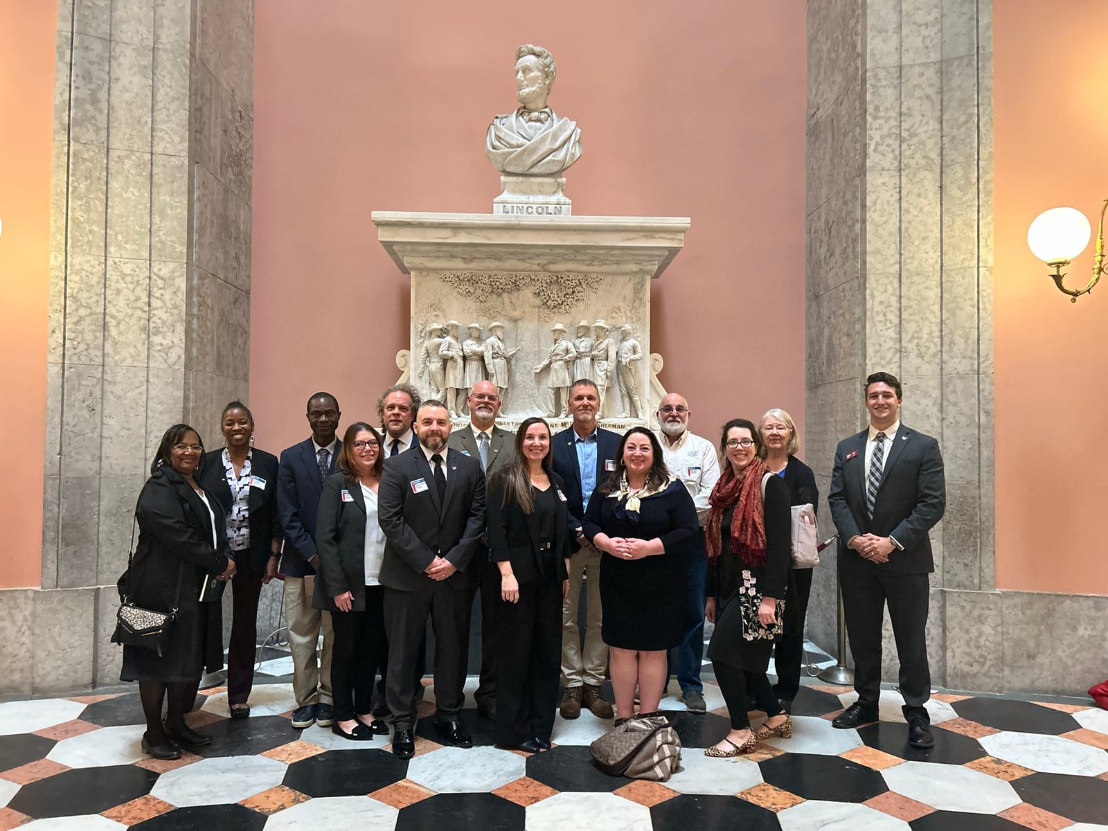 A group of people posing inside a marble hall beneath a bust statue.